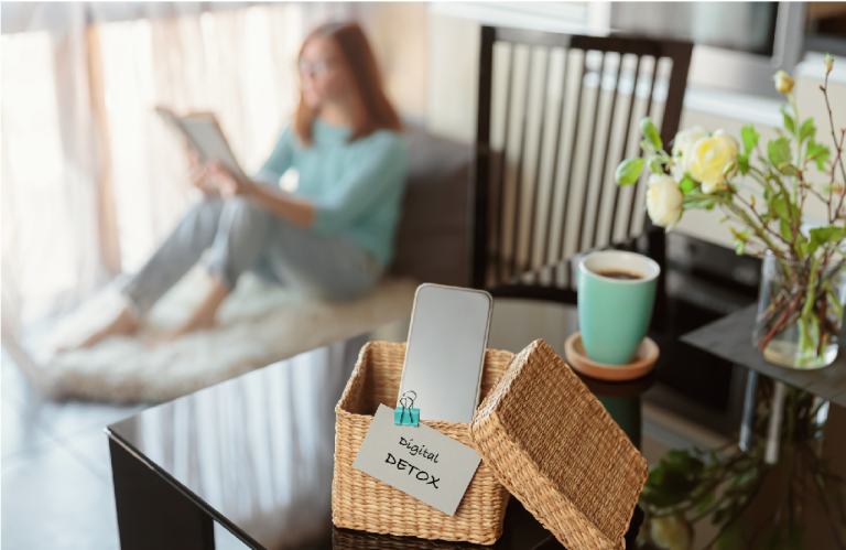 The image depicts a peaceful scene of a woman enjoying a digital detox in her living room. The focus is on a small, open woven box placed on a sleek black table. Inside the box lies a smartphone, face down, symbolizing a conscious break from the digital world. A small handwritten note clipped to the box proclaims "Digital Detox," emphasizing the woman's intention to disconnect and unwind. In the soft-focus background, the woman sits comfortably on a plush white rug near a large window, bathed in natural light. She is engrossed in a book, her attention fully captured by the physical pages rather than a screen. A steaming cup of coffee rests on the table beside the box, suggesting a cozy and relaxing atmosphere. A vase with delicate white flowers adds a touch of serenity to the scene. The overall composition of the image, with the sharp focus on the "Digital Detox" box and the blurred background, effectively conveys the essence of disconnecting from technology and embracing the present moment. The woman's relaxed posture and the calming elements of the room further reinforce the theme of tranquility and mindful living.